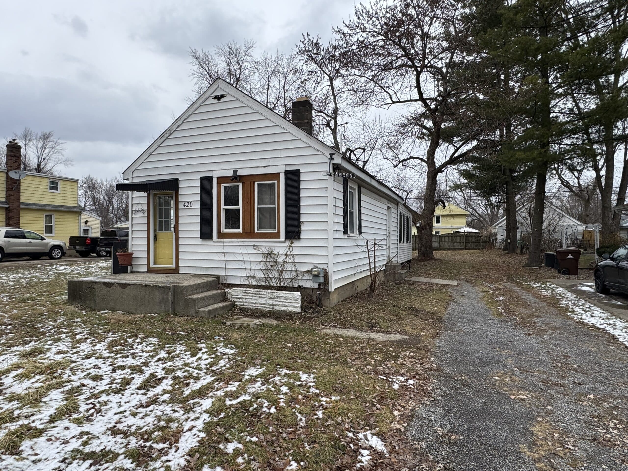 Small white house with a yellow front door and gravel driveway in DeWitt